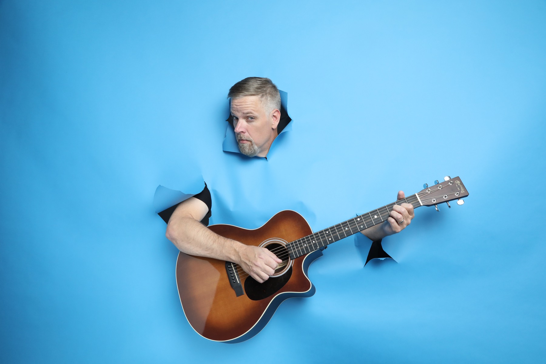 Jonnie W. bursting through blue paper holding an acoustic guitar